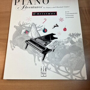 Może przedstawiać: Książka zatytułowana "Piano Adventures Christmas" autorstwa Nancy i Randalla Fabera. Książka jest dla starszych początkujących i zawiera czytanie z nut do wypełniania skarpet. Okładka przedstawia czarne pianino ze skrzydłami, renifera i sanie.