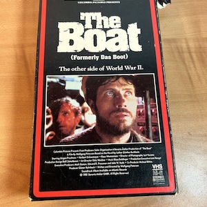 May include: A VHS tape case for the film "The Boat" (Formerly Das Boot). The case is black with red trim and features a photo of the film's star, Jürgen Prochnow, looking intently at the camera. The text on the case reads "Columbia Pictures Presents The Boat (Formerly Das Boot) The other side of World War II."