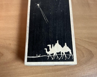 May include: A black and white woodblock print of three camels with riders traveling under a starry night sky with a bright star in the top left corner.