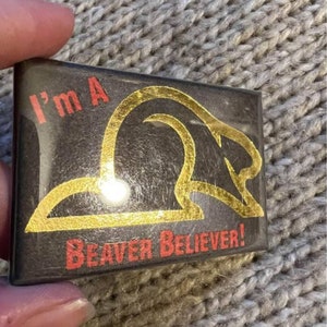 Puede incluir: Un botón negro con letras doradas que dice "I'm a Beaver Believer!" con una silueta de castor dorada.
