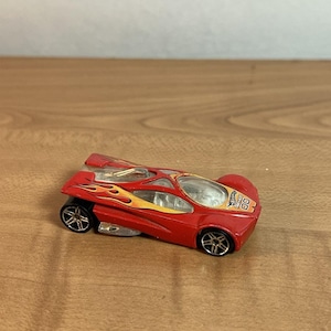 May include: A red die-cast toy car featuring yellow and orange flame graphics along the sides, a clear plastic cockpit canopy, black wheels with silver rims, and the Hot Wheels logo printed on the front bonnet with the number 00.