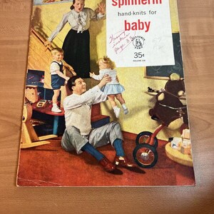 以下が含まれることがあります： 4人家族が写っているビンテージ雑誌の表紙。表紙には「spinnerin hand-knits for baby」というタイトルと、35セントの価格が記載されています。雑誌は108巻です。表紙の画像には、母親、父親、そして2人の子供が写っています。父親は床に座って、子供を膝に乗せています。母親は背景に立って、もう一人の子供を抱いています。家族は、ロッキングホース、トライシクル、人形などのおもちゃに囲まれています。