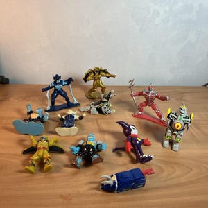 Puede incluir: Una colección de figuras de acción coloridas, que incluyen robots, superhéroes y otros personajes de fantasía. Las figuras están en varias poses y presentan colores como oro, azul, rojo y amarillo. Los juguetes están colocados sobre una superficie de madera.