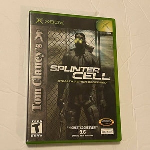 May include: A green Xbox game case for Tom Clancy's Splinter Cell, a stealth action game. The cover features a man in black clothing with night vision goggles, standing in front of a metal fence. The game is rated T for Teen by the ESRB. The case includes the text "HIGHEST SCORE EVER!" and "9.6 - OFFICIAL XBOX MAGAZINE".
