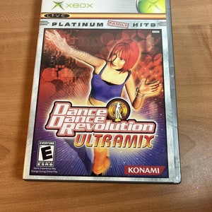 May include: Xbox game case for Dance Dance Revolution Ultramix. The case is silver with a red, orange, and purple background. The game features a woman in a purple and orange outfit dancing. The ESRB rating is Everyone.