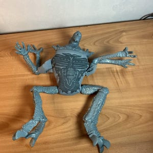 May include: A gray, articulated action figure of a creature with a detailed torso, multiple arms, and legs. The figure has a textured surface and is posed with arms outstretched and legs bent, giving it a dynamic appearance.