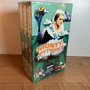 Peut inclure: Un coffret DVD de Monty Python's Flying Circus. La boîte est verte avec des images d'une femme couronnée, de nuages et d'autres graphiques. L'ensemble contient plusieurs disques.