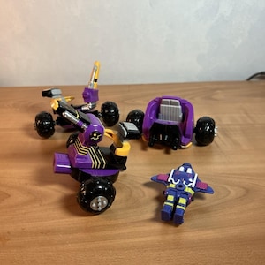May include: A collection of purple and black toy vehicles, including a three-wheeled ATV, a buggy, and a small robot figure. The toys have yellow and silver accents and black wheels, set against a wooden surface.