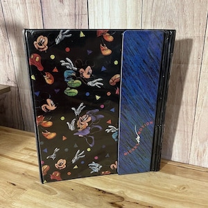 Peut inclure: Un livre noir avec un motif Mickey Mouse présentant diverses poses et des accents colorés. La tranche a un design bleu et noir avec le texte "Mickey in Motion". Le livre est sur une surface en bois.
