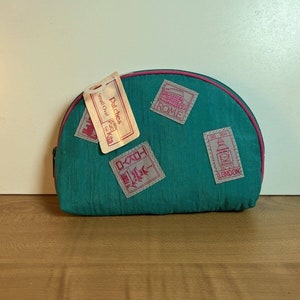 May include: Teal blue oval-shaped zippered pouch with a pink trim. The pouch has several fabric patches with images of landmarks from around the world, including Rome, Tokyo, and London. The pouch has a tag attached that reads "Pal's Ches Small Oval Pac Kits".
