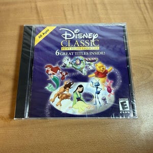 Puede incluir: Un CD-ROM sellado de Disney Classic Print Studio Collection. La portada presenta personajes de varias películas de Disney, incluyendo a Ariel, Buzz Lightyear y Winnie the Pooh. El texto dice "6 Great Titles Inside!" y "All Ages".