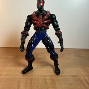 May include: A black action figure with a red spider-like design on the chest and head, and blue accents on the legs. The figure is standing on a wooden surface.