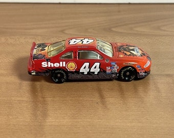 May include: A red, white, and black toy race car with the number 44 on the side. The car is sponsored by Shell and has a black and white graphic of a wolf on the side. The car is on a wooden surface.