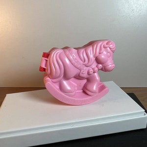 Può includere: Un giocattolo di cavallo a dondolo in plastica rosa con una sella rossa e un'imbragatura bianca e rosa. Il cavallo ha una criniera e una coda in plastica rosa.