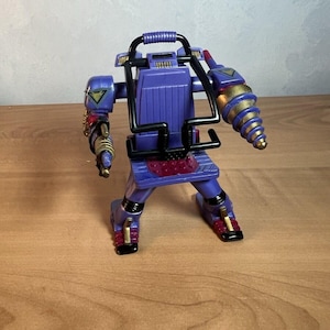 May include: A purple and gold robot toy with a drill arm and a black handle on the back. The robot has a red and purple skirt and black boots.