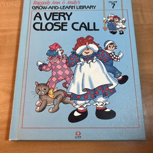 Op de afbeelding: Een blauwe boekomslag met de titel "A Very Close Call" uit de Grow-and-Learn Library van Raggedy Ann & Andy. De omslag toont illustraties van Raggedy Ann en Andy, een clown en een kat.