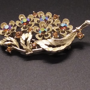 May include: A gold-toned brooch with a floral design. The brooch features several small flowers with iridescent rhinestones in the center. The flowers are surrounded by leaves and a single leaf extends to the side.