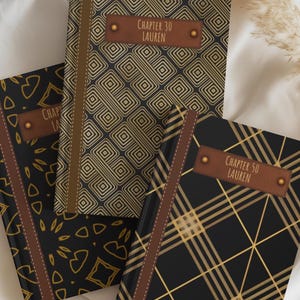 May include: Three hardback journals with black and gold geometric patterns. Each features a brown spine and a rectangular brown label with gold studs. The labels read Chapter 30 Lauren, Chapter 50 Lauren, and one partially visible label.