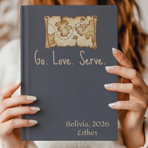 Könnte beinhalten: Ein dunkelgraues Notizbuch mit festem Einband und einer Aquarellillustration einer Schatzkarte oben. Der Text "Go. Love. Serve." ist mittig aufgedruckt, während "Bolivia, 2026" und der Name "Esther" unten in hellem Beige stehen.