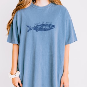 May include: A blue t-shirt with a graphic of a fish and the words "Fishes and Loaves" in a dark blue color. The shirt has short sleeves and a crew neck.