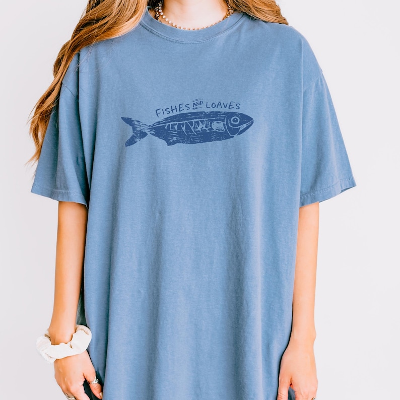 May include: A blue t-shirt with a graphic of a fish and the words "Fishes and Loaves" in a dark blue color. The shirt has short sleeves and a crew neck.