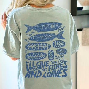 May include: A light blue t-shirt with a blue graphic of two fish, leaves, and the text "I'll give you all of my fishes and loaves." The design is printed on the back of the shirt.