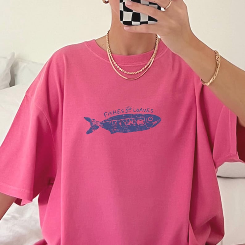 May include: A pink t-shirt with a blue illustration of a fish and the words "Fishes & Loaves". The shirt has short sleeves and a crew neck. The shirt is made of a soft material.