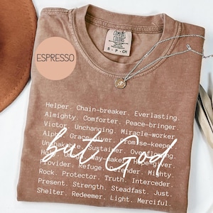 May include: A brown t-shirt with the words "But God" in white text. The shirt also has a list of words describing God, including "Helper", "Chain-breaker", "Everlasting", "Almighty", "Comforter", "Peace-bringer", "Victor", "Unchanging", "Miracle-worker", "Alpha", "Grace giver", "Promise-keeper", "Unshakable", "Sustainer", "Overflowing", "Holy", "Omega", "Way-maker", "Hope-giver", "Provider", "Refuge", "Defender", "Mighty", "Rock", "Protector", "Truth", "Interceder", "Present", "Strength", "Steadfast", "Just", "Shelter", "Redeemer", "Light", and "Merciful".