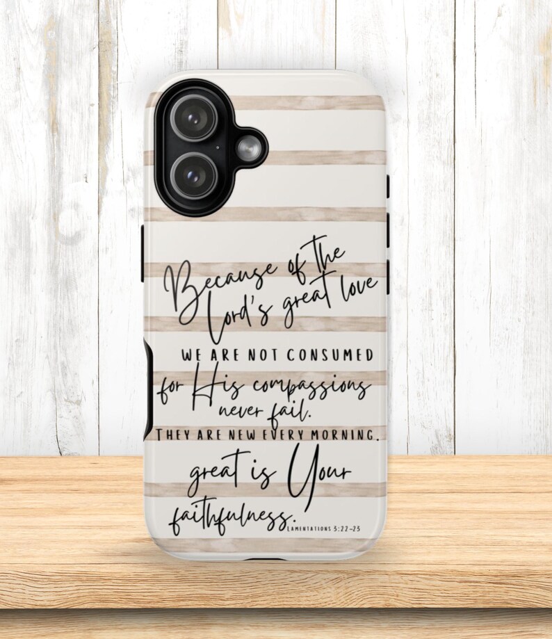 May include: A white and beige striped phone case with the text "Because of the Lord's great love...great is Your faithfulness." The case has a black camera cutout and is on a wooden surface.