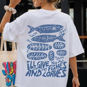 May include: White t-shirt with a blue fish and leaf design. The text "I'LL GIVE YOU ALL OF MY FISHES AND LOAVES" is printed in blue. The shirt is a casual, oversized style.