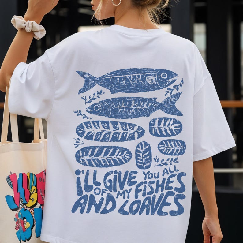 May include: White t-shirt with a blue fish and leaf design. The text "I'LL GIVE YOU ALL OF MY FISHES AND LOAVES" is printed in blue. The shirt is a casual, oversized style.