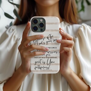 May include: A white and beige striped phone case with the text "Because of the Lord's great love...great is Your faithfulness." The case is held in a person's hands. The phone case is designed to fit a smartphone.