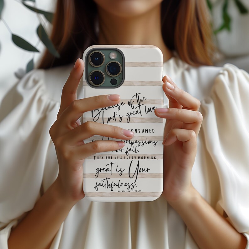 May include: A white and beige striped phone case with the text "Because of the Lord's great love...great is Your faithfulness." The case is held in a person's hands. The phone case is designed to fit a smartphone.