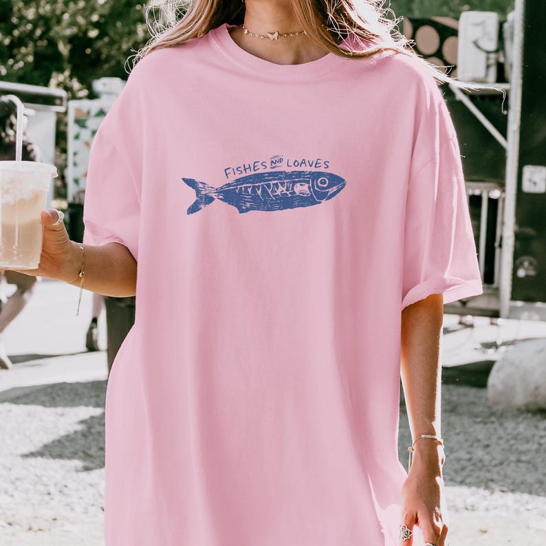 May include: A light pink t-shirt featuring a blue fish illustration with the words "Fishes and Loaves" printed above it. The shirt has short sleeves and a relaxed fit.