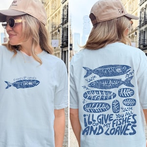 May include: Light blue t-shirt with a navy blue fish graphic and the text "Fishes & Loaves" on the front. The back of the shirt features two fish, loaves, and the text "I'll give you all of my fishes and loaves."