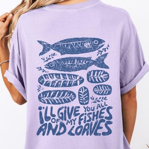 May include: A light purple t-shirt featuring a blue graphic of two fish, leaves, and loaves of bread. The text "I'll give you all of my fishes and loaves" is printed in blue at the bottom of the shirt.