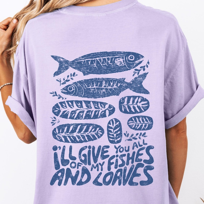 May include: A light purple t-shirt featuring a blue graphic of two fish, leaves, and loaves of bread. The text "I'll give you all of my fishes and loaves" is printed in blue at the bottom of the shirt.