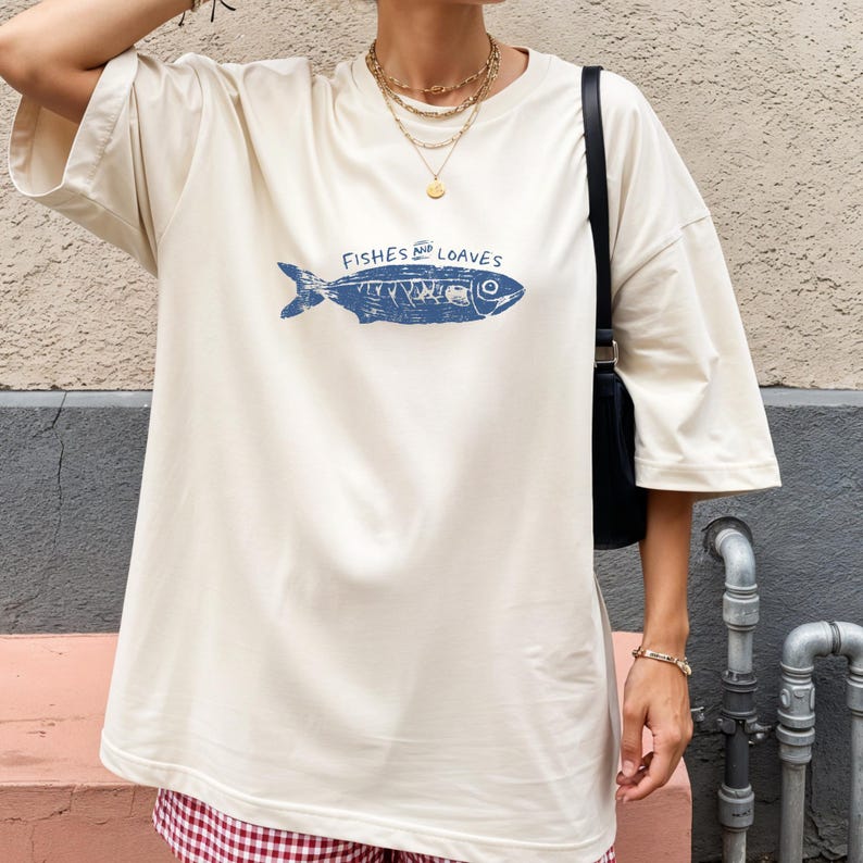 May include: Cream-colored t-shirt with a blue fish graphic and the words "Fishes and Loaves." The shirt has short sleeves and a relaxed fit. The bottom of the shirt is partially visible, paired with a red and white checkered skirt.