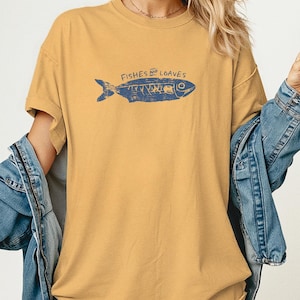 May include: Mustard yellow t-shirt with a blue illustration of a fish and the words "Fishes and Loaves". The short-sleeved shirt is made of a soft material and has a relaxed fit.