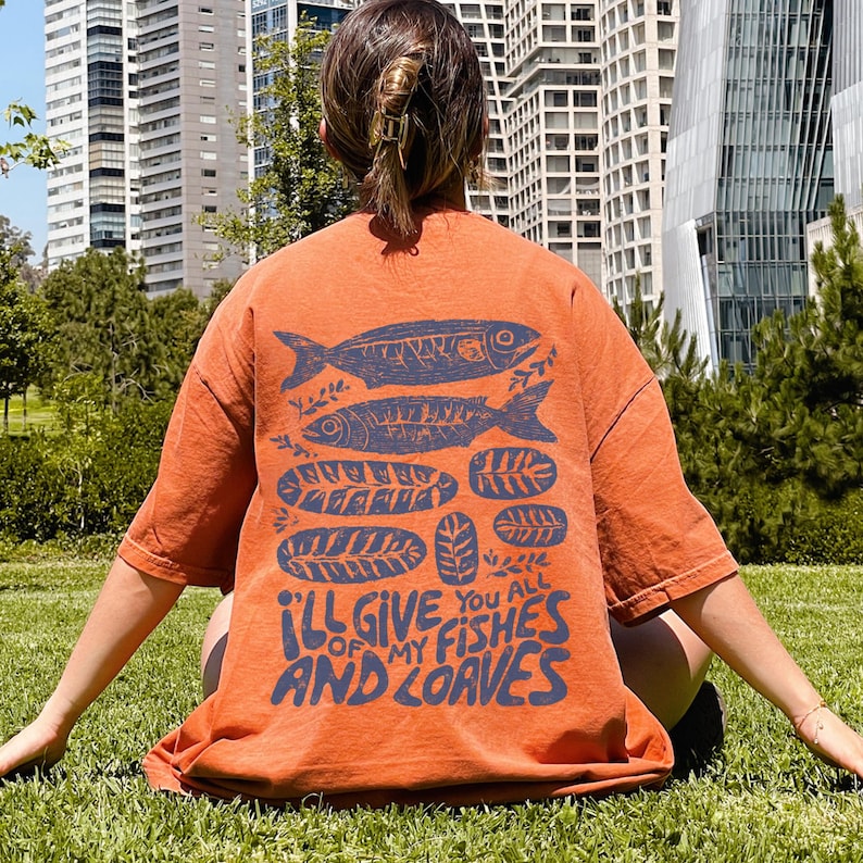 May include: Burnt orange t-shirt with a blue graphic of fish and leaves. The text "I'll give you all of my fishes and loaves" is printed below the graphic. The shirt is oversized and has short sleeves.