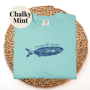 May include: A light blue t-shirt with a navy blue fish graphic and the words "Fishes and Loaves". The shirt is on a woven basket. The words "Chalky Mint" are in a circle in the upper left corner.