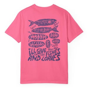 May include: A pink t-shirt with a purple graphic design on the back. The design includes two fish, several loaves of bread, and the text "I'll give you all of my fishes and loaves."
