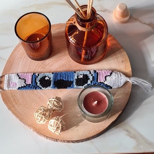 Puede incluir: Un brazalete de amistad azul y rosa con un diseño de personaje de dibujos animados. El brazalete está sobre una superficie de madera con una vela pequeña y una botella de aceite esencial.