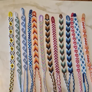 May include: A collection of 14 colourful friendship bracelets with various patterns, including chevron, diamond, and swirl designs. The bracelets are made with colourful embroidery floss and have knotted ends.