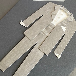 May include: A gray paper card with the text "Congratulations and Best Wishes" on a white label. A gray paper suit is cut out and layered on the card. The suit has a white shirt with a black bow tie.