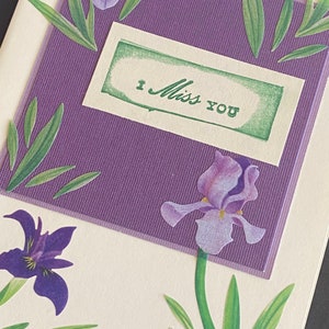 May include: A purple and white card with a green rectangle that says "I Miss You". The card is decorated with purple and green flowers.