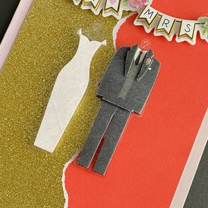 May include: A wedding card with a gold glitter background and a red background. The card features a white dress and a black tuxedo with a bow tie. The card also features a banner that says "Mr. & Mrs."
