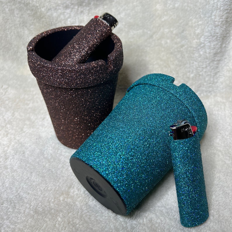 May include: Two glitter ashtrays, one brown and one teal, with matching lighters. The lighters are made of metal and have a red accent.