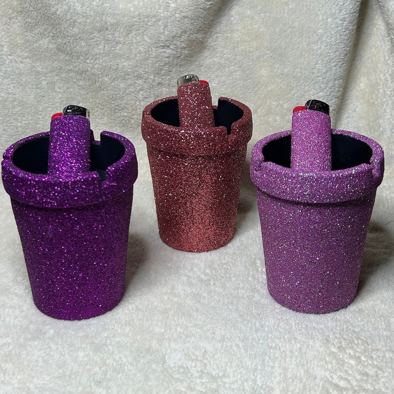 May include: Three glitter-covered ashtrays in different colors: purple, pink, and brown. Each ashtray has a small, black lighter inside.