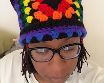 May include: A colorful, handmade crochet hat with cat ears. The hat features a rainbow pattern in the center, surrounded by black and purple. The hat is worn by a person wearing glasses and a white t-shirt.